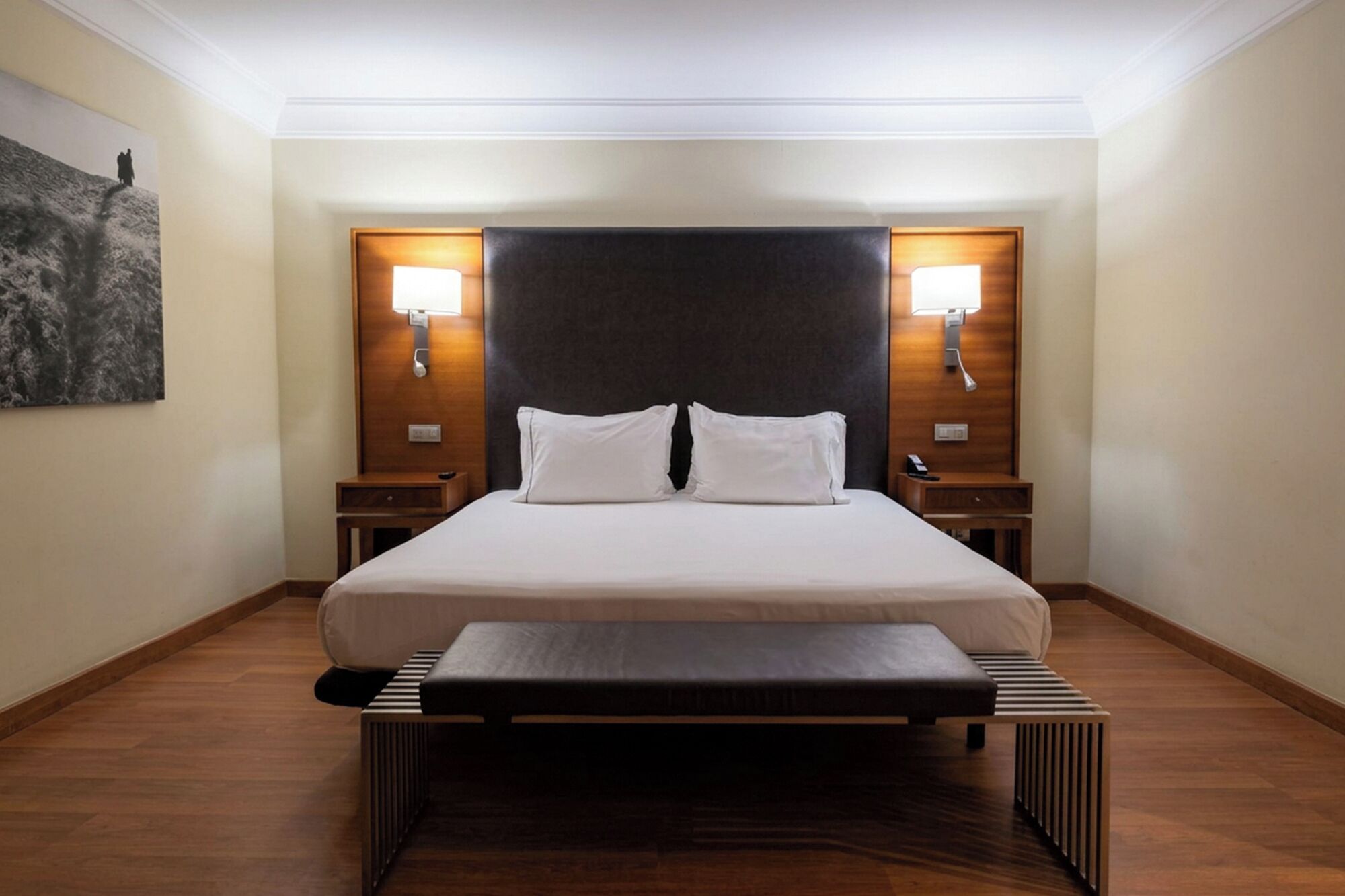 Habitación de hotel con una cama grande, dos mesitas de noche, un banco al pie de la cama y una obra de arte colgada en la pared.
