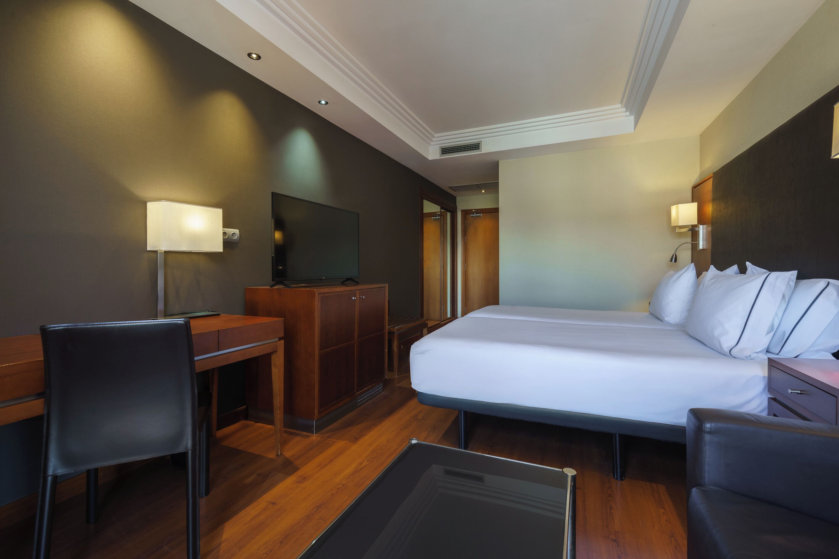 A hotel room with a bed, desk, chair, TV, wardrobe, and bedside table.