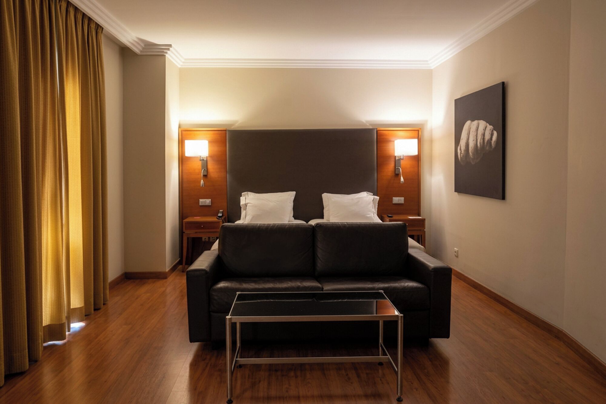 Habitación de hotel con sofá de cuero oscuro, una mesita pequeña, dos lámparas de noche y un cuadro enmarcado en la pared.