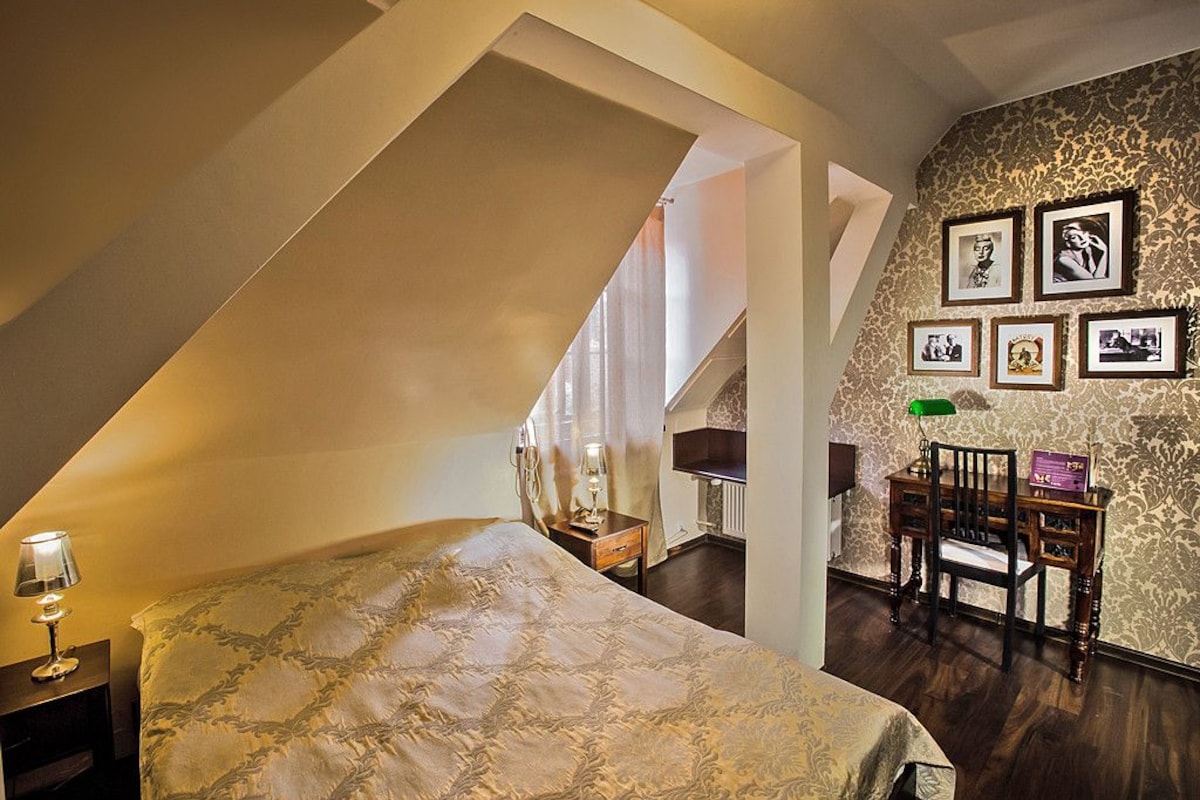 A bedroom with a sloped ceiling, a bed with a patterned bedspread, a desk with a chair, and a wall with framed pictures.
