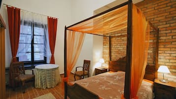 A four-poster bed with a canopy, a round table, two chairs, a nightstand with a lamp, and a brick wall.