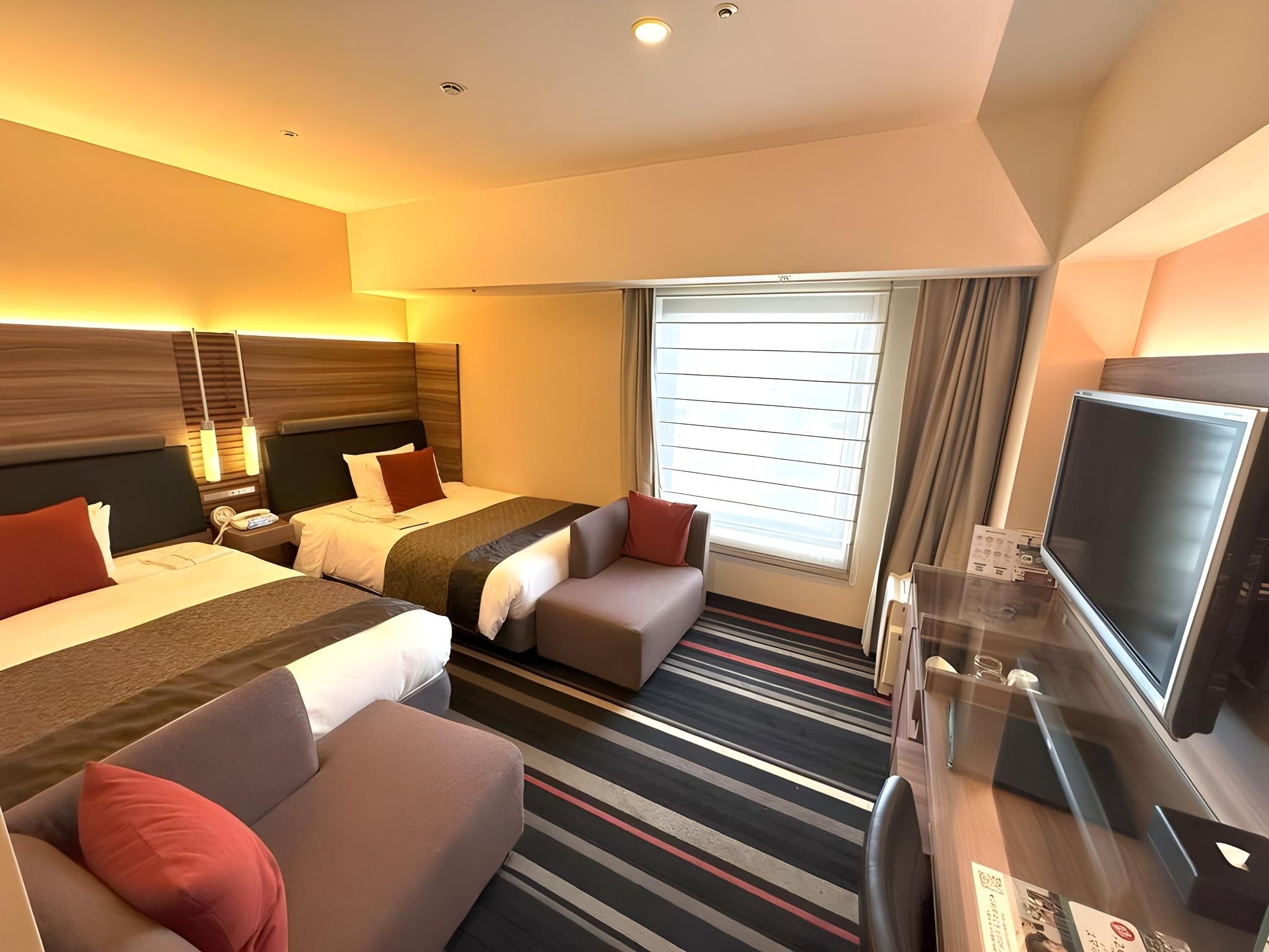 A hotel room with two beds, a sofa, a TV, and a window with blinds.