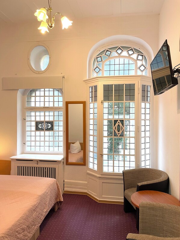 A bedroom with a bed, a window with decorative glass, a mirror, and a television mounted on the wall. - Hotel Castell (Berlin)
