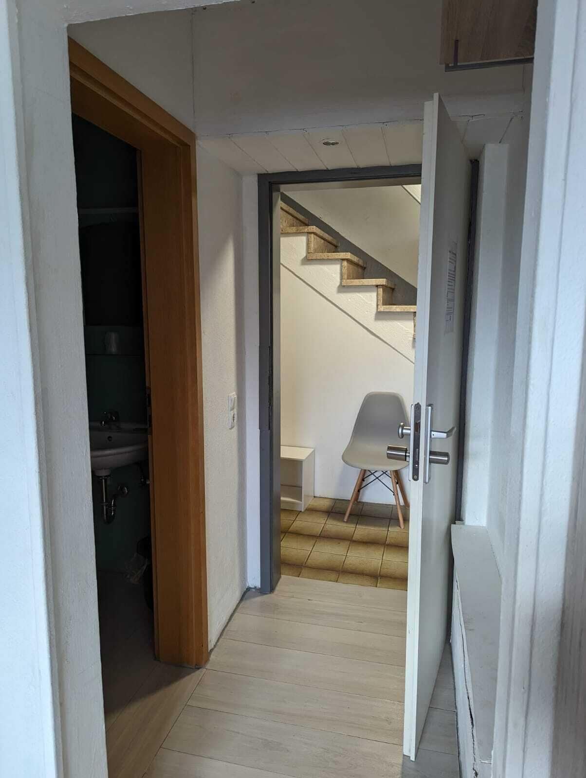 A hallway with a wooden door leading to a bathroom, a white chair, and a staircase.