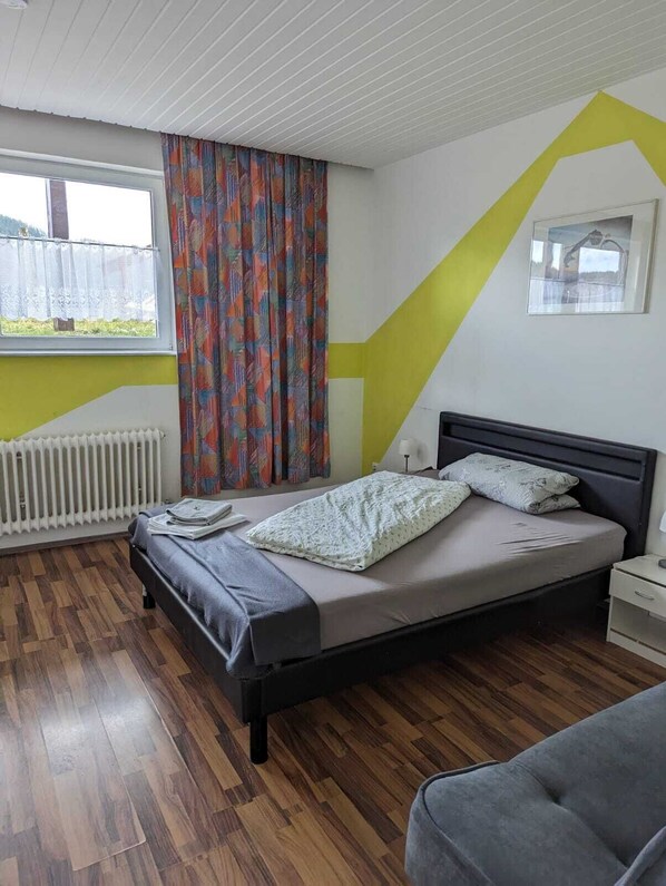 A bedroom with a bed, a nightstand, a window with curtains, a radiator, and a wooden floor.
