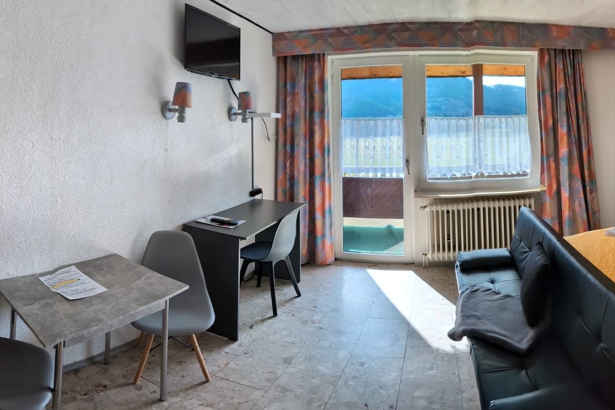 A bedroom with a bed, a desk, and a chair. There is a window with curtains and a radiator below it.