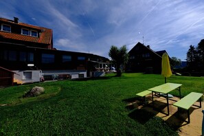 Uma área externa com grama, mesas para piquenique e um guarda-sol, um edifício com telhado vermelho e um céu claro.