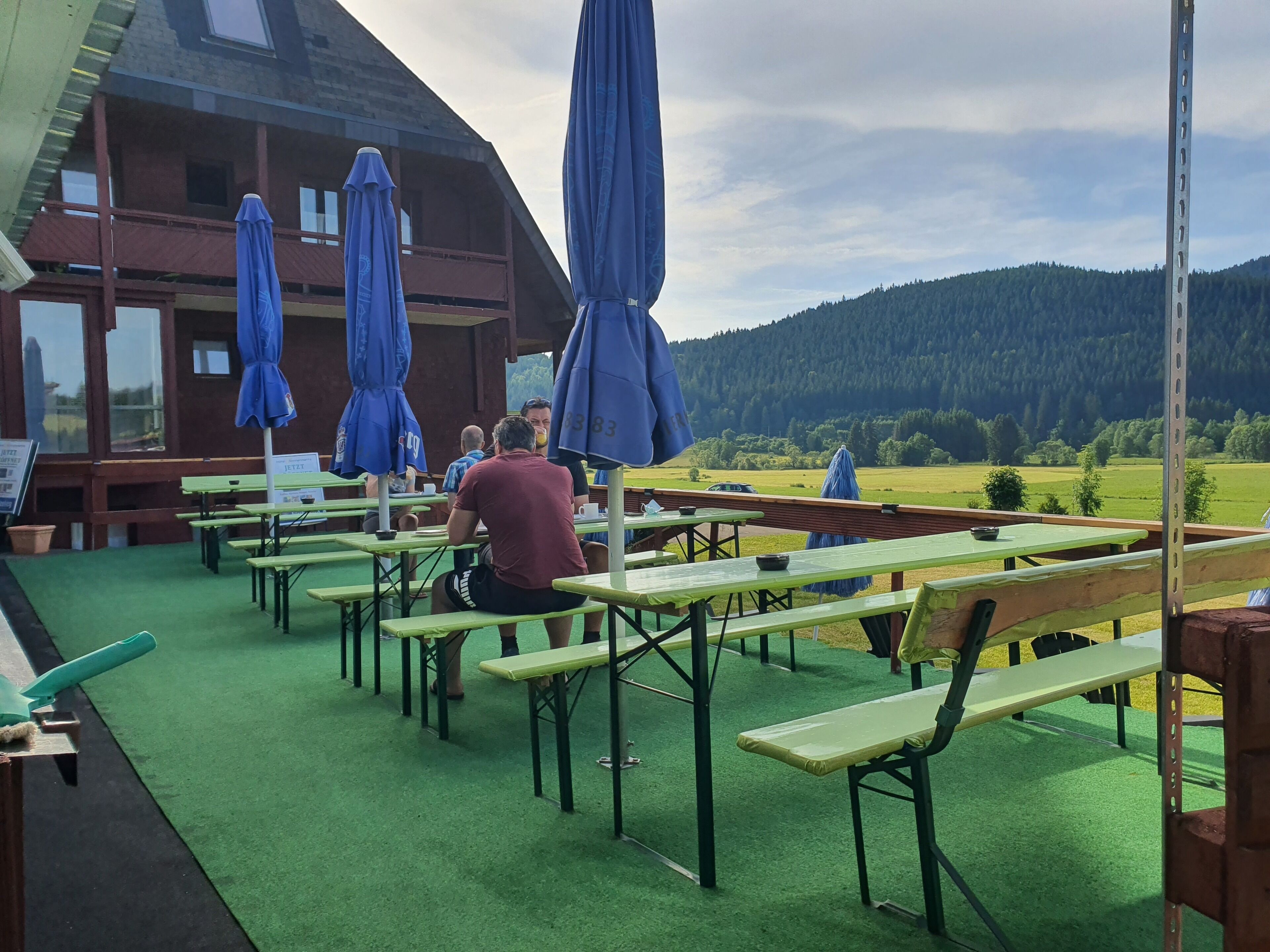 An outdoor seating area with green tables and benches, blue umbrellas, and a view of a forested hill.
