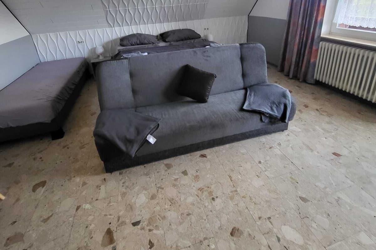 A room with a grey sofa, a patterned ceiling, a window with curtains, and a radiator.