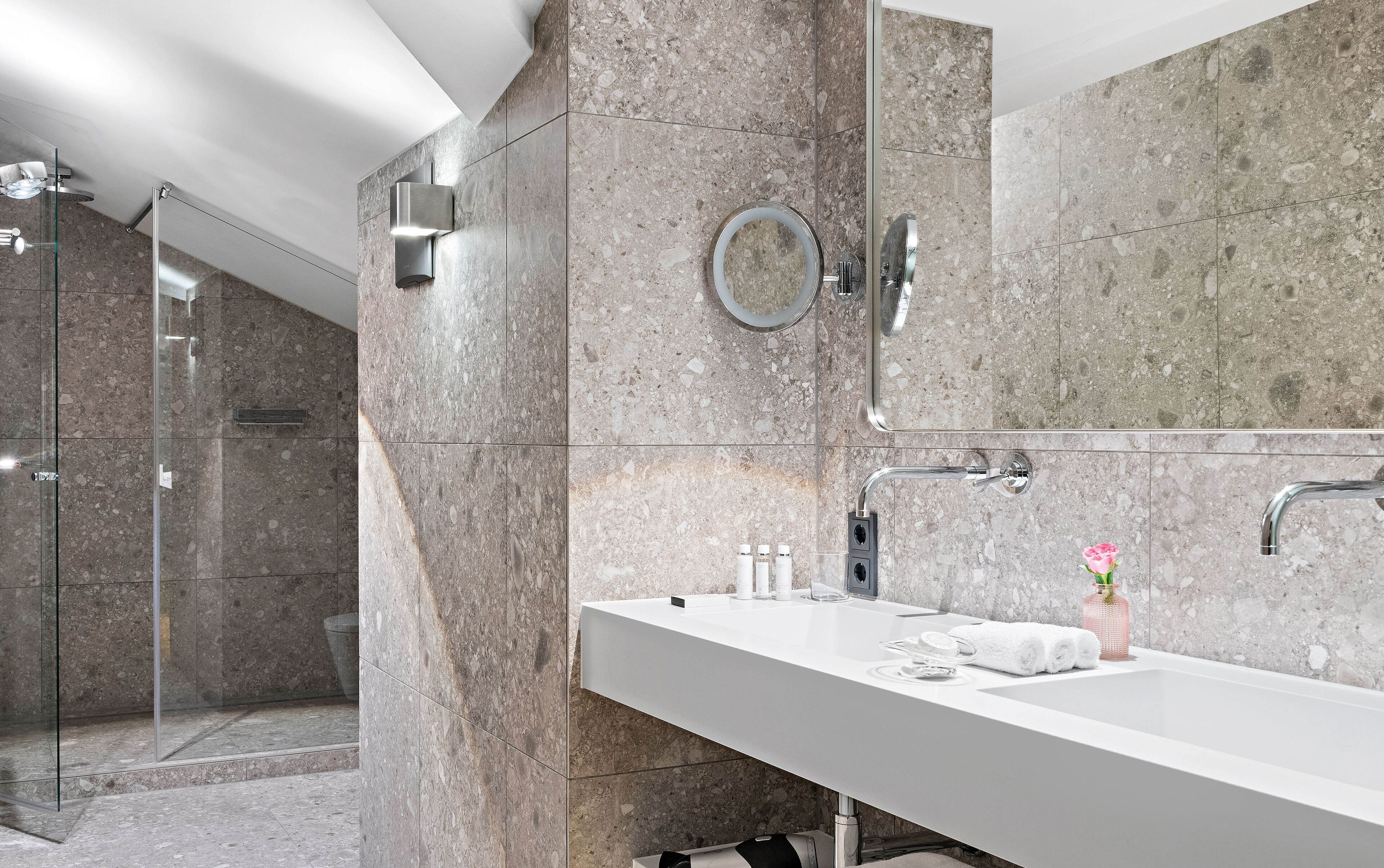 A modern bathroom with a glass-enclosed shower, a large mirror, and a white vanity with toiletries.