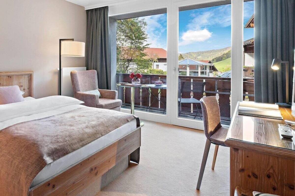 A hotel room with a bed, two chairs, a desk, and a balcony with a view.