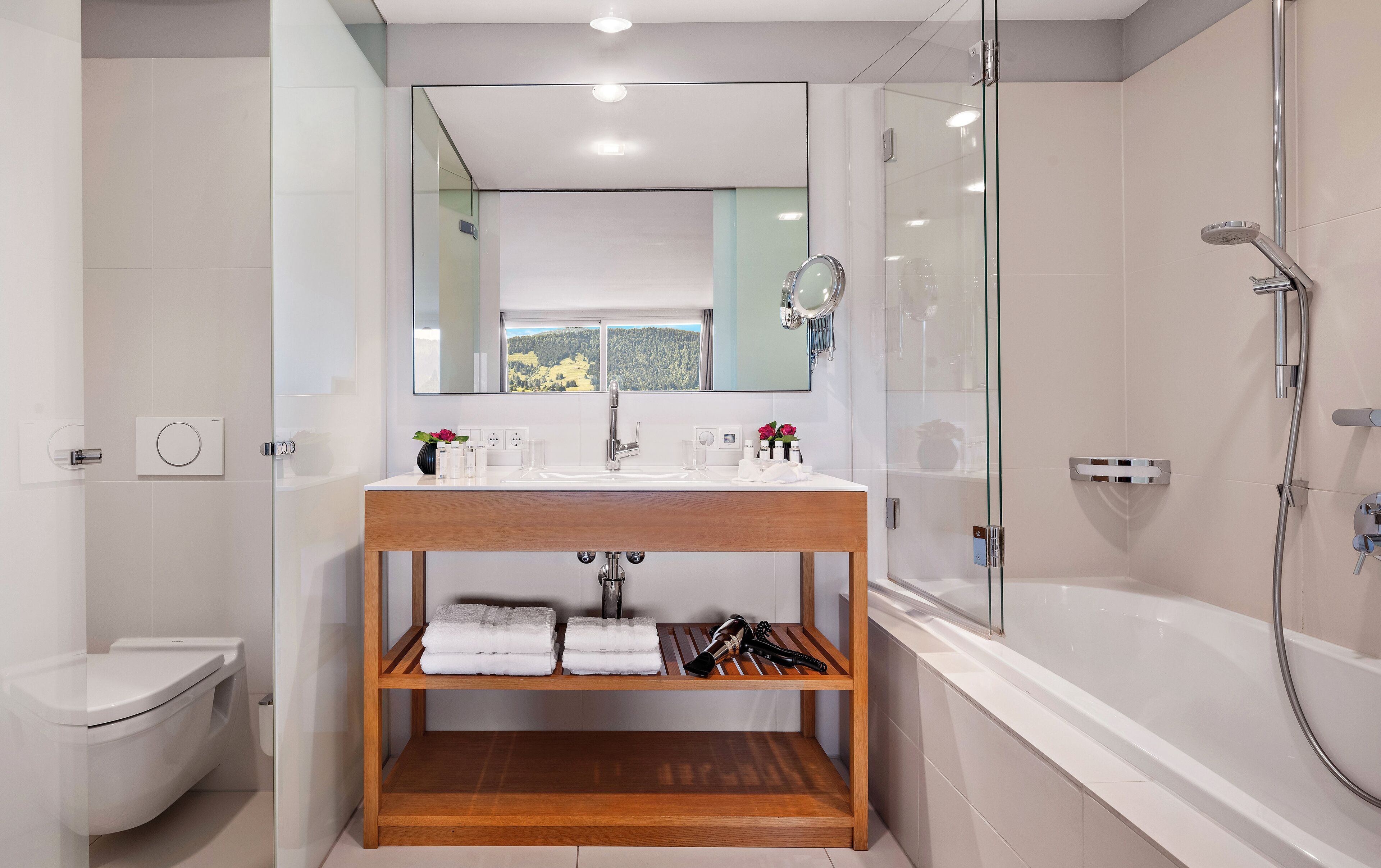 Un baño moderno con ducha envuelta en vidrio, tocador de madera, inodoro y vista al exterior a través de una ventana.