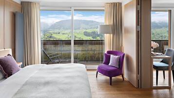 Habitación de hotel con una cama grande, un sillón púrpura, una mesita con una silla y un balcón con vistas a colinas verdes.