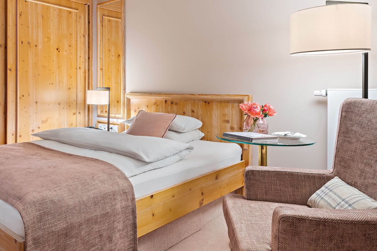 A hotel room with a wooden bed, a glass side table with a book and flowers, a lamp, and a chair.