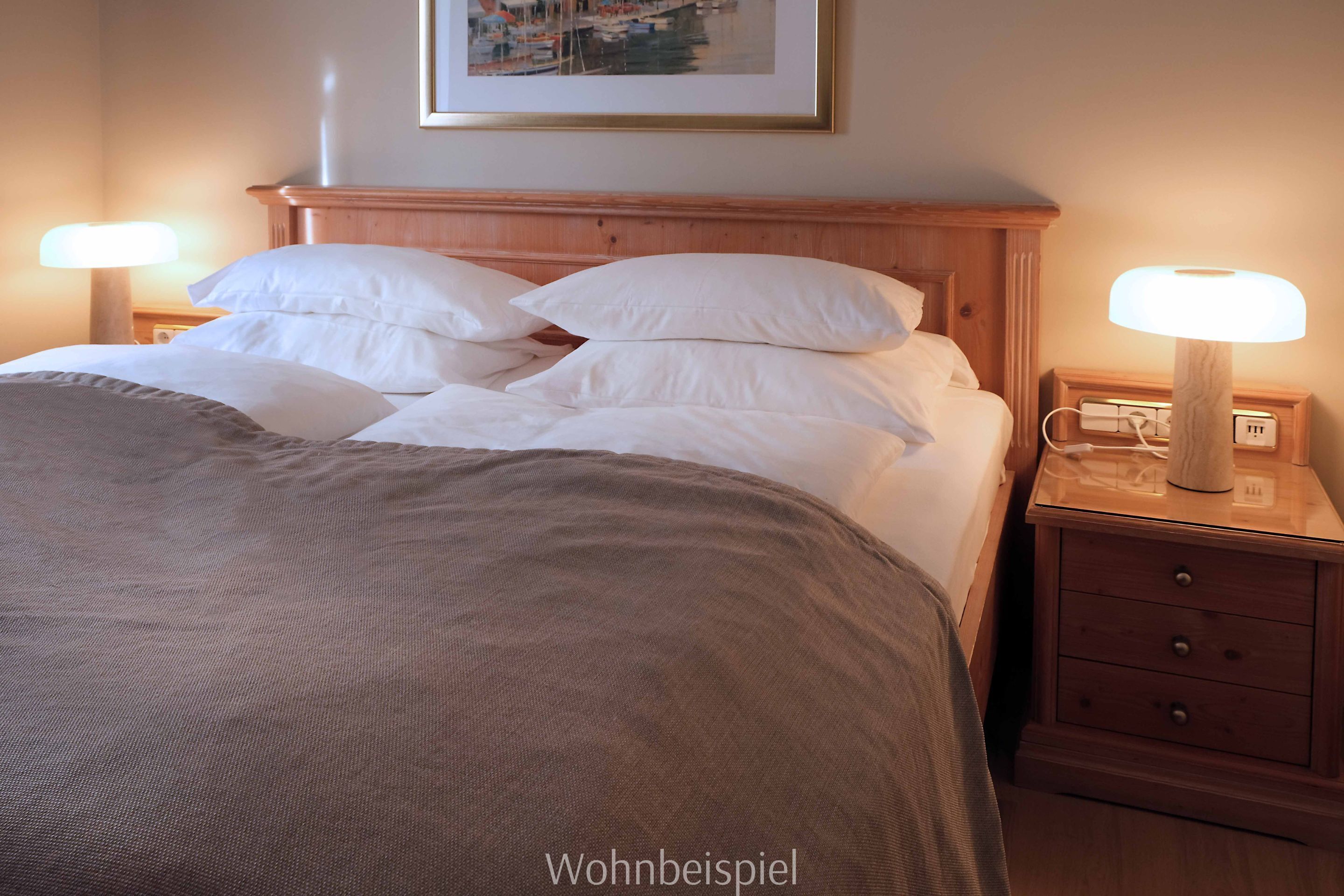 Un lit bien fait, avec du linge de lit blanc et une couverture marron, flanqué de deux tables de chevet sur lesquelles sont posées des lampes, dont l’une est équipée d’une multiprise.