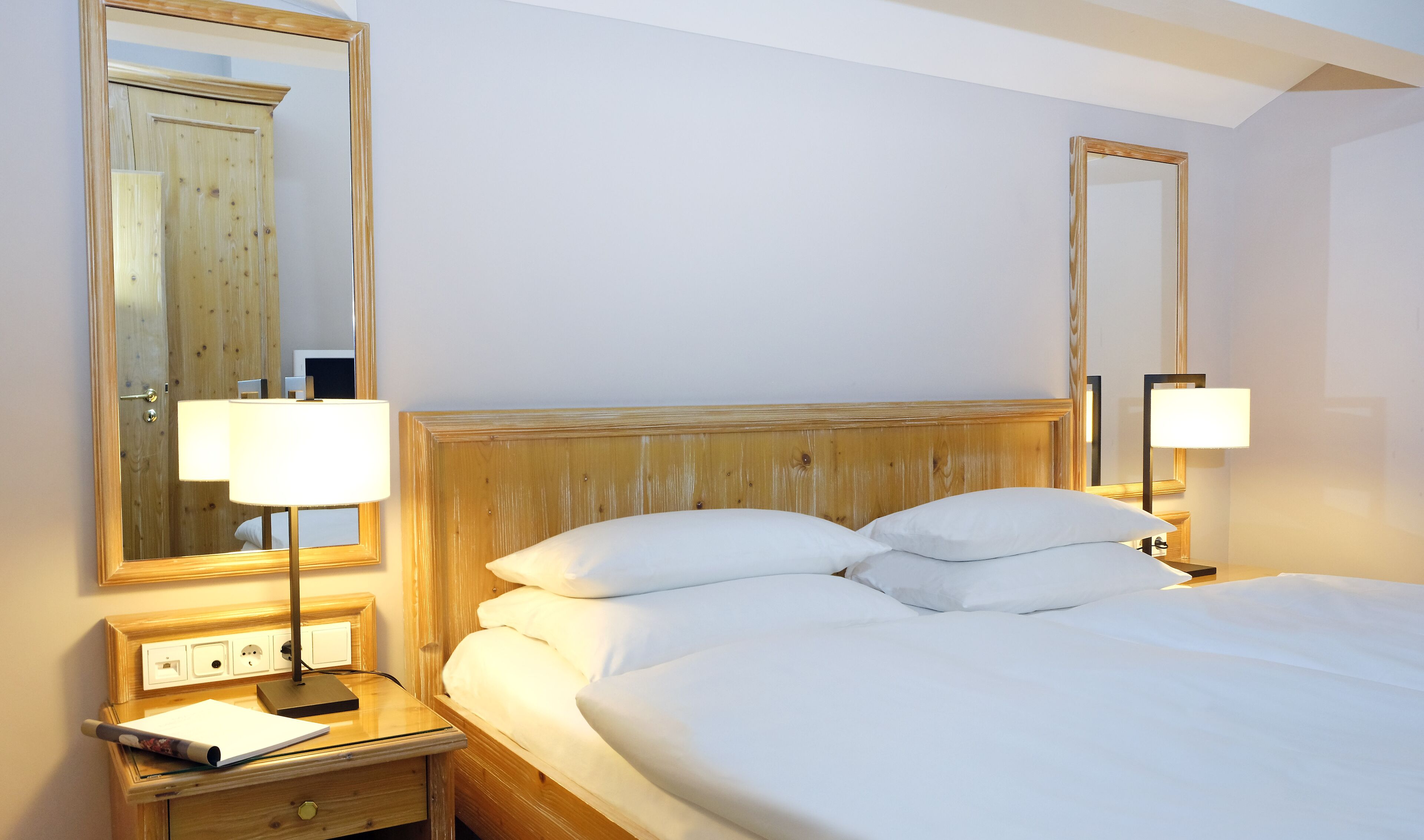 A hotel room with a wooden headboard, two bedside lamps, a nightstand with a lamp and a notepad, and a large mirror on the wall.