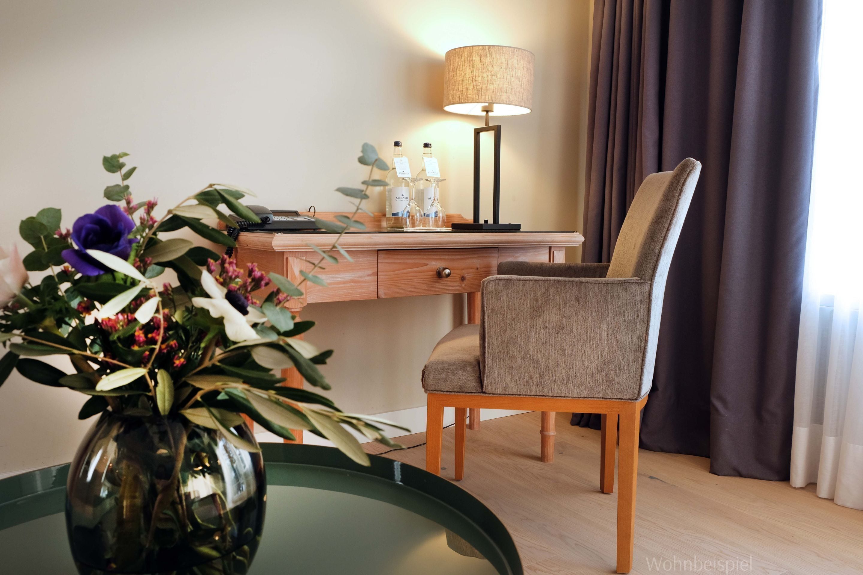 Een hotelkamer met een houten bureau, een stoel, een lamp, een vaas met bloemen en uitzicht door gordijnen.