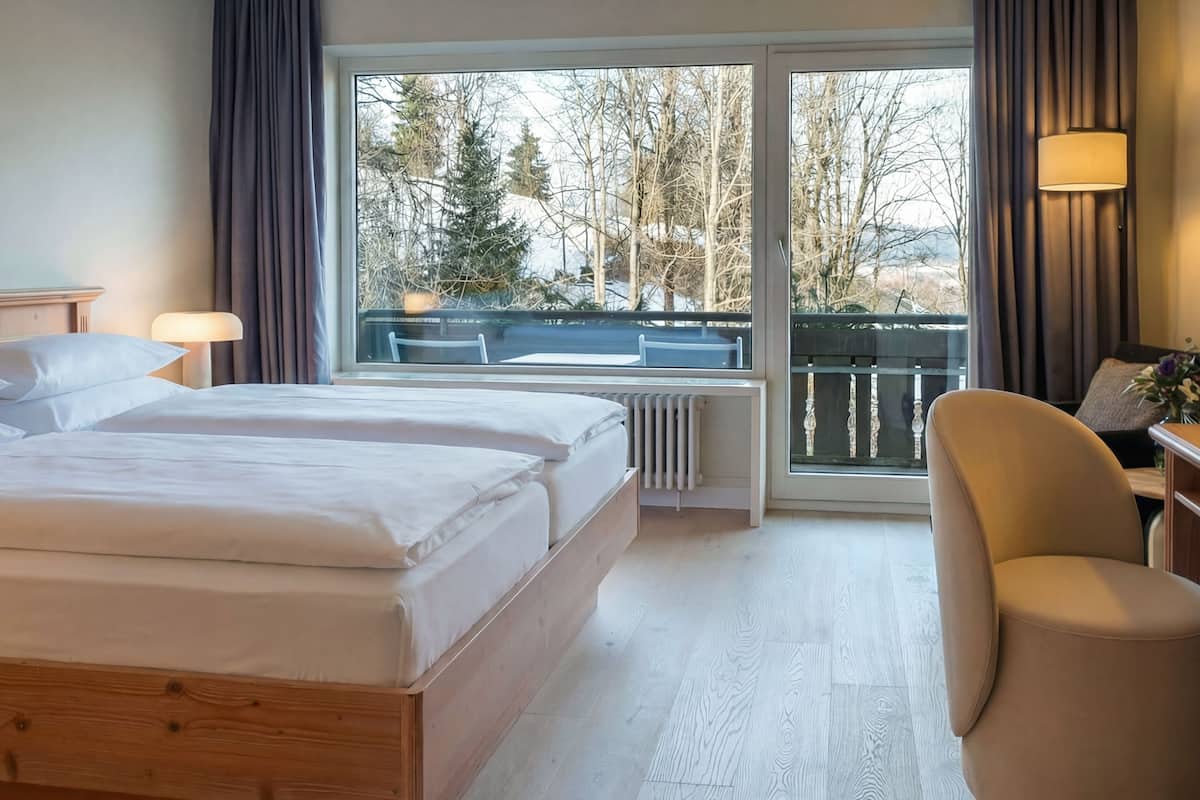 A hotel room with a large bed, a chair, a table, and a view of trees and a balcony.