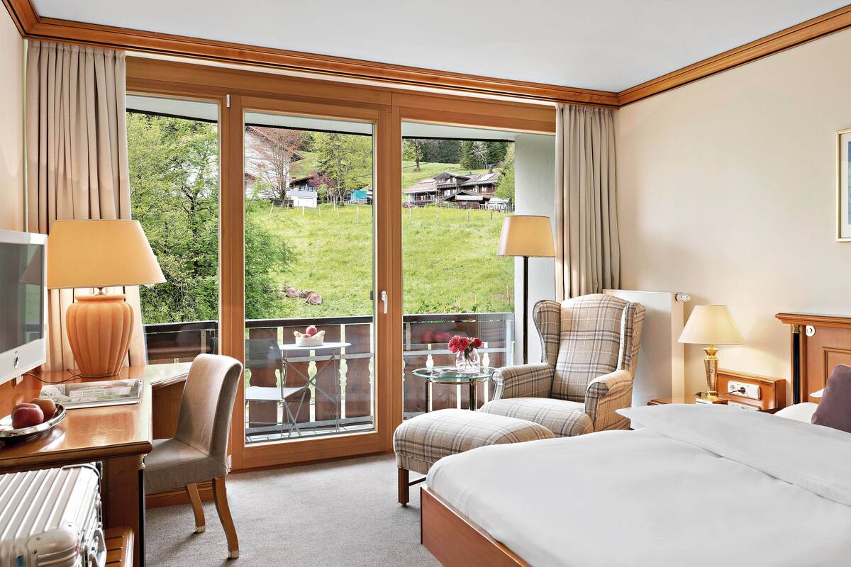 A hotel room with a bed, two chairs, a desk with a lamp, a TV, and a balcony with a view of a green landscape.
