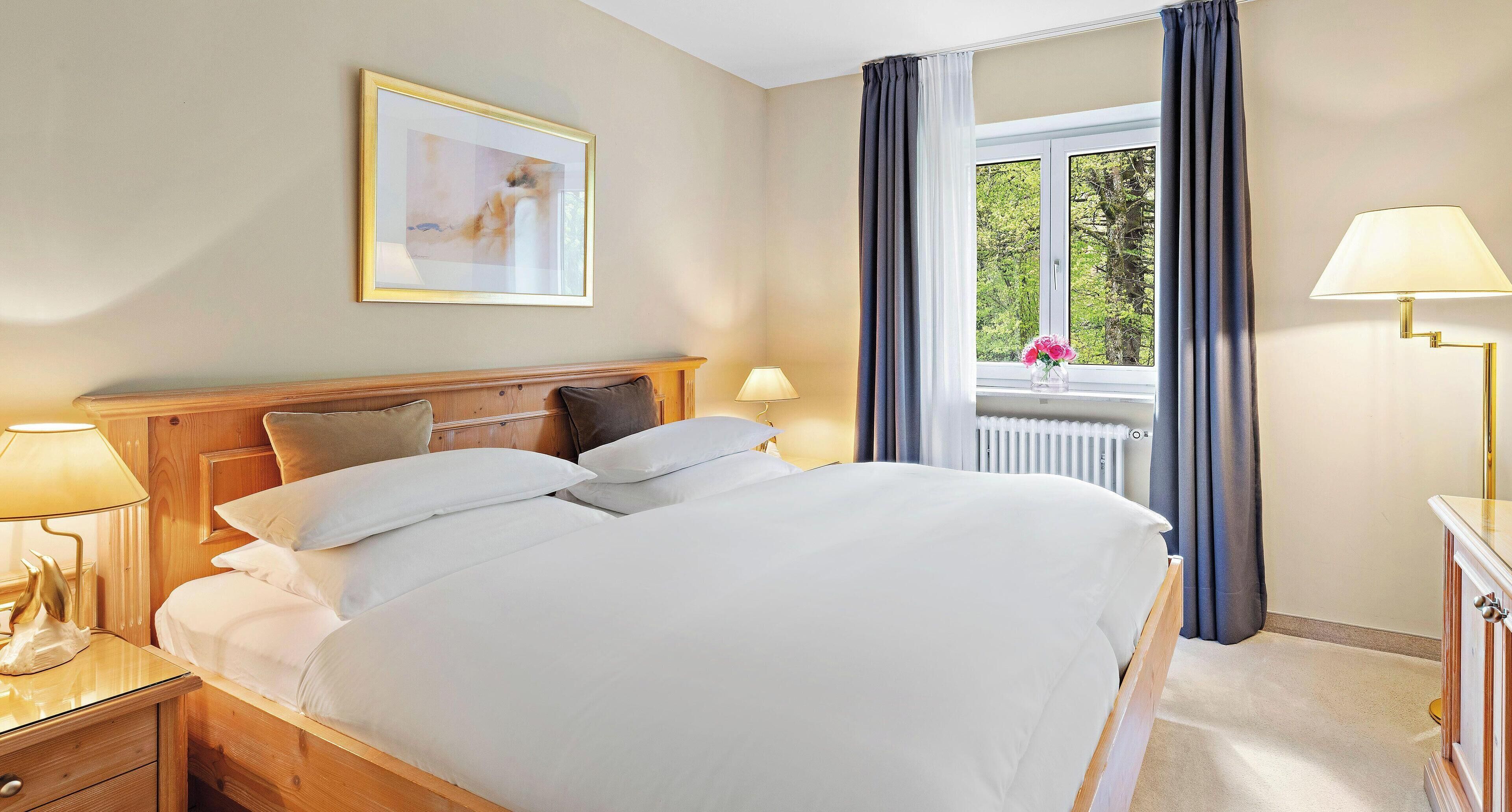 A neatly made bed with white linens, a wooden headboard, and a bedside table with a lamp. A window with curtains and a view of trees is visible, and a framed picture hangs on the wall.