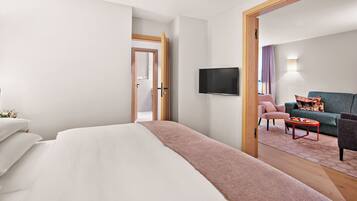 Ein modernes Hotelzimmer mit einem großen Bett, einem flachbildfernseher an der Wand, einer Sitzecke mit Sofa und Sesseln sowie einer Holztür, die in einen anderen Raum führt.