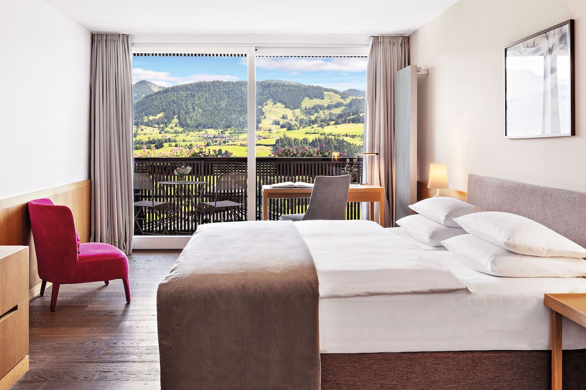 A hotel room with a large bed, a desk, a chair, and a view of a green landscape through a balcony.