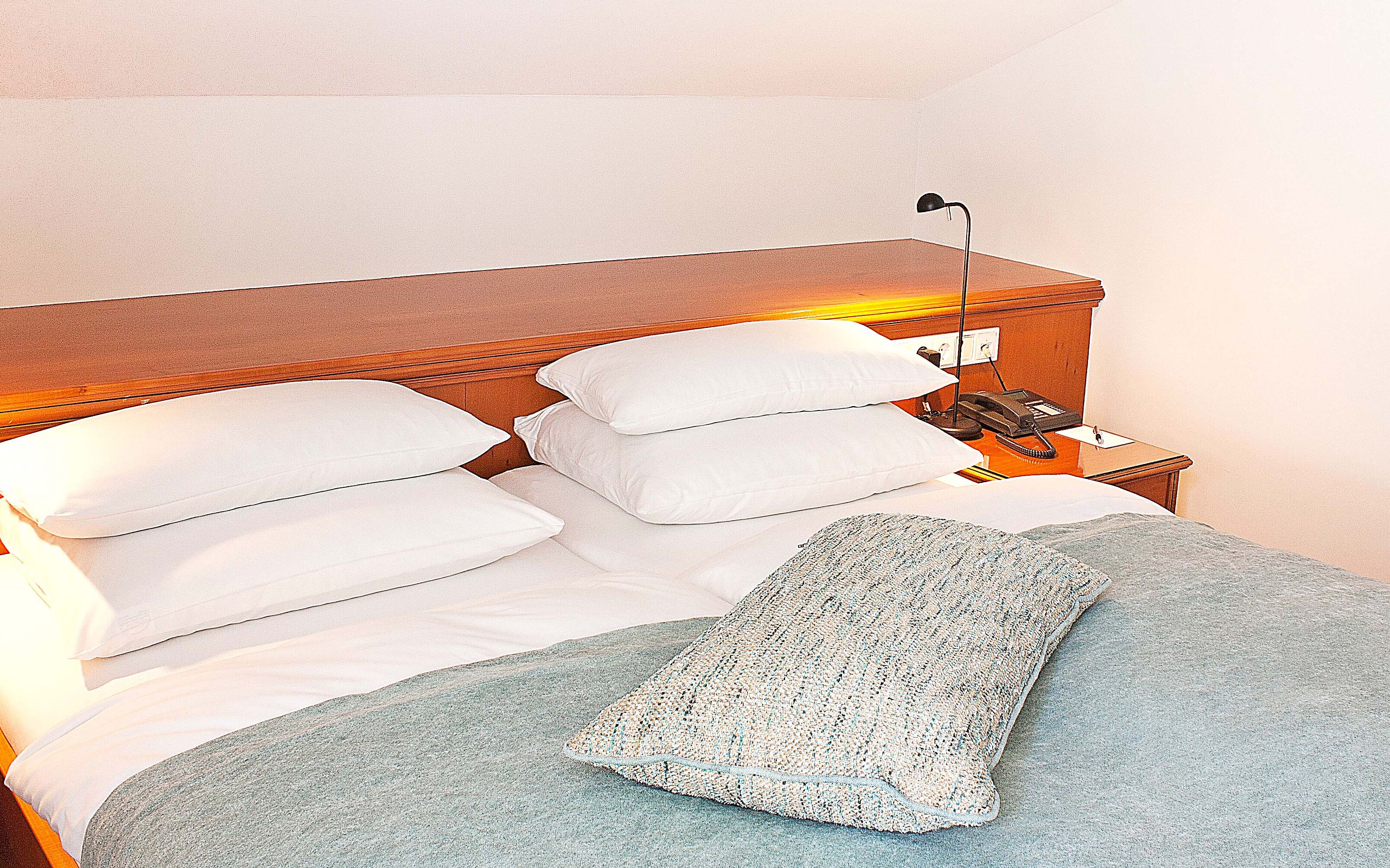 Una cama bien tendida con almohadas blancas y una manta gris, una lámpara de noche y un teléfono sobre una mesita de madera.