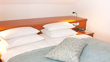 Una cama bien tendida con almohadas blancas y una manta gris, una lámpara de noche y un teléfono sobre una mesita de madera.