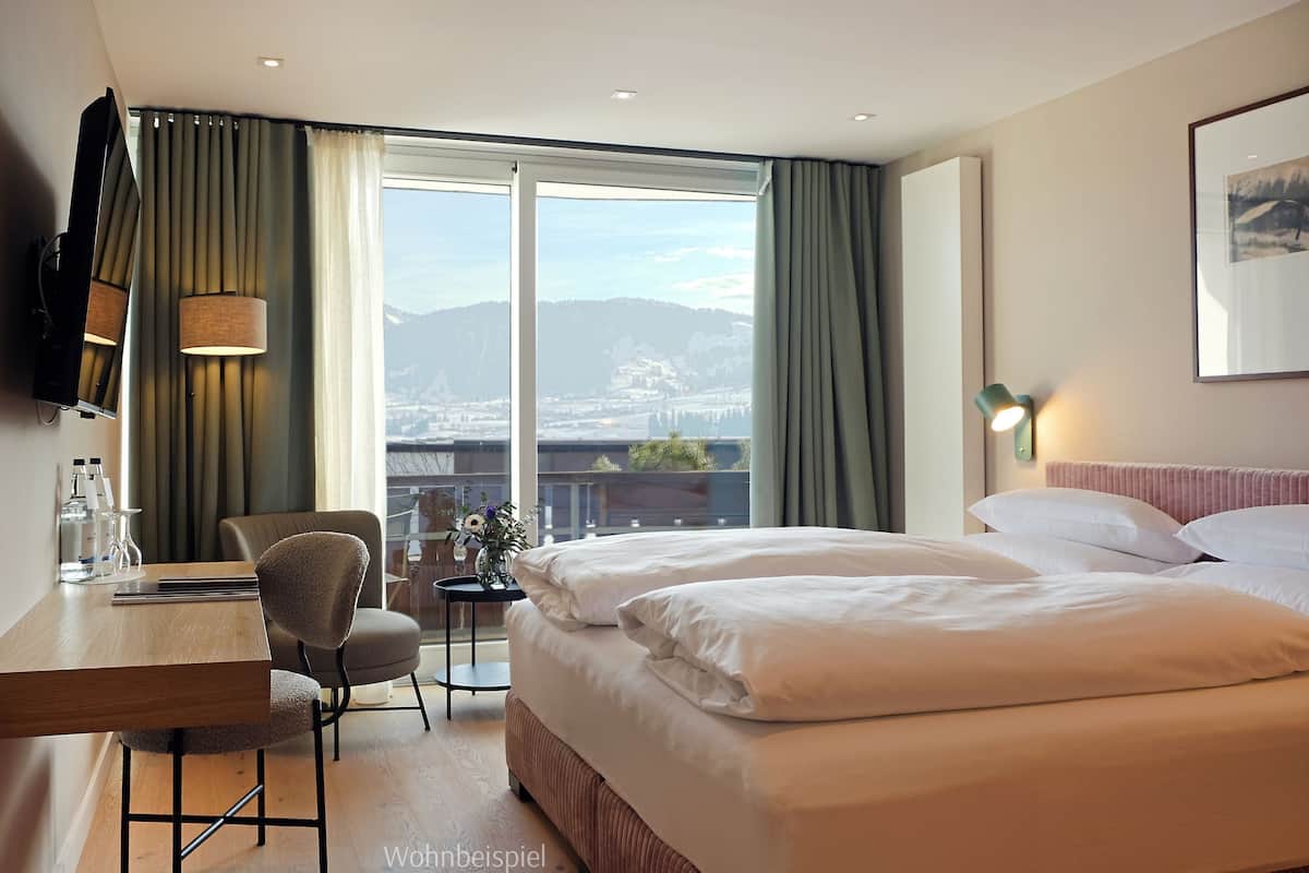 A hotel room with a large bed, a desk with a chair, a TV mounted on the wall, and a view of a cityscape through the window.