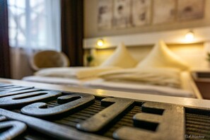 Een detailfoto van een bed met witte lakens en kussens, een nachtkastje met een lamp, en een raam met gordijnen.
