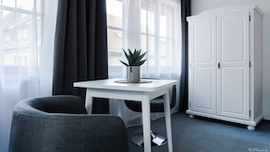 Una habitación con una mesa blanca, una silla gris, un armario blanco y una ventana con cortinas traslúcidas.