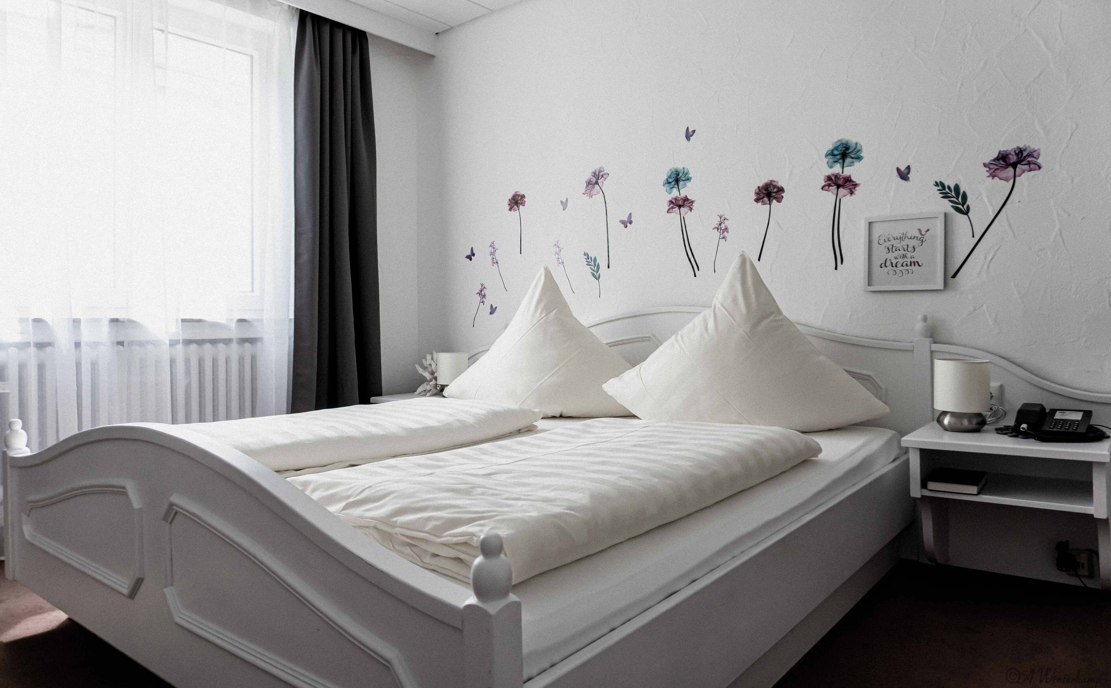 A neatly made bed with white linens and pillows, a bedside table with a lamp and phone, and a wall decorated with floral decals.