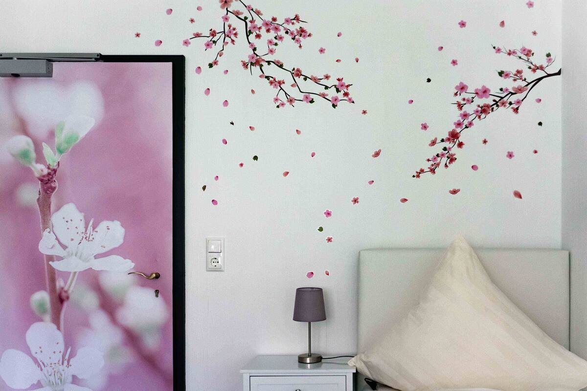 Una habitación con una puerta decorada con motivos florales, una mesita de noche con una lámpara, y una pared adornada con una rama y flores de cerezo.