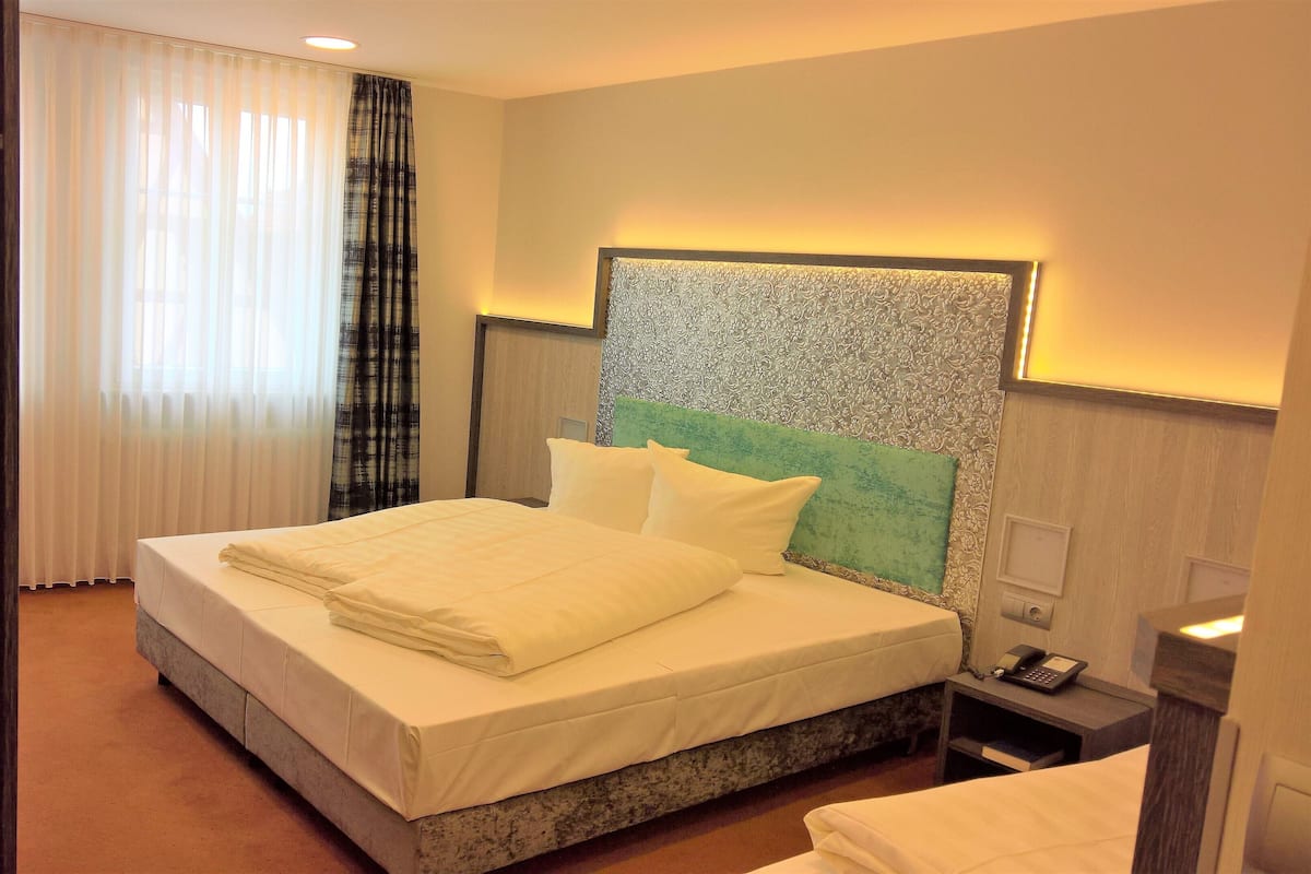 Habitación de hotel con una cama grande, cabecera con diseño estampado, mesita de noche con teléfono y ventana con cortinas traslúcidas.