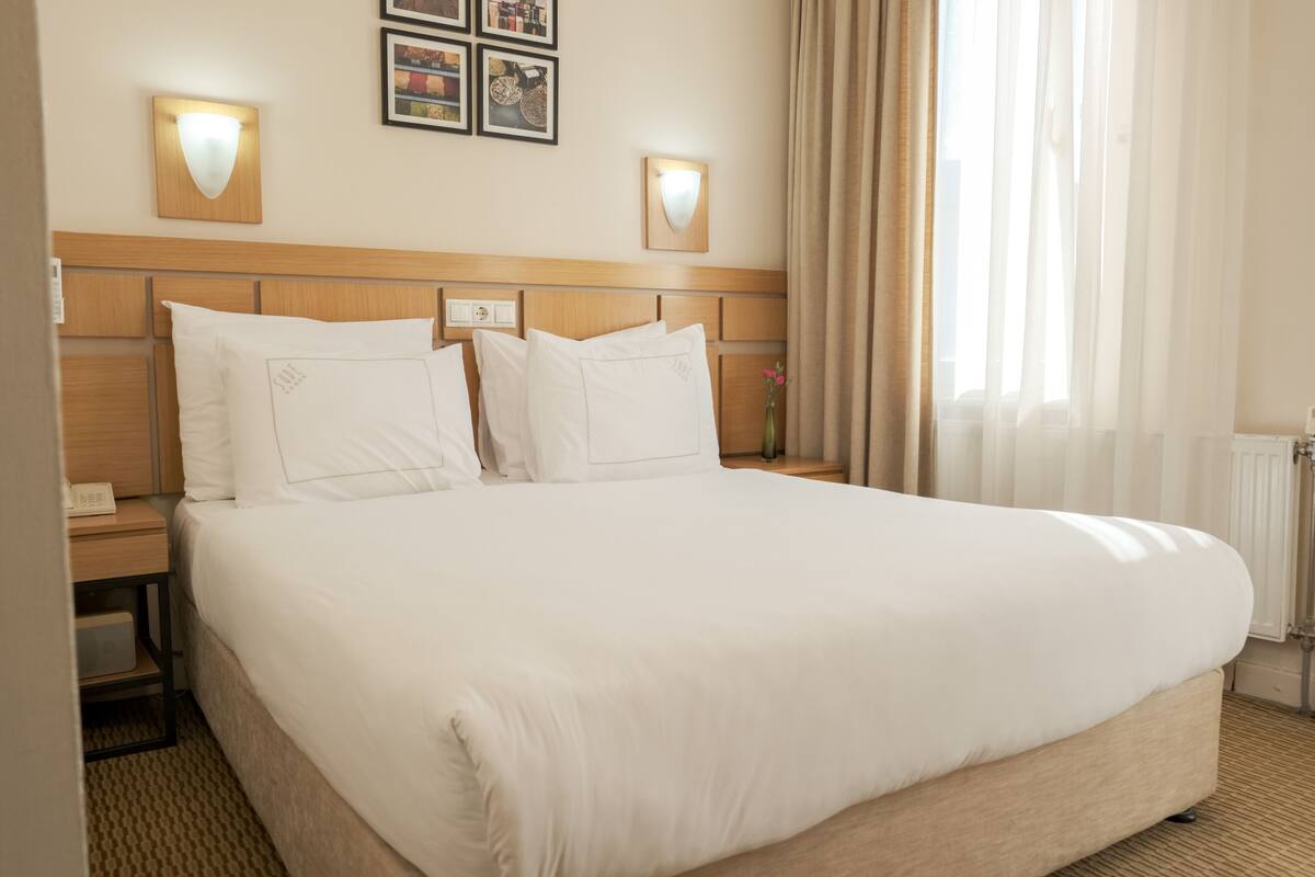 A hotel room with a large bed, two bedside tables, a nightstand, a lamp, and a window with curtains.