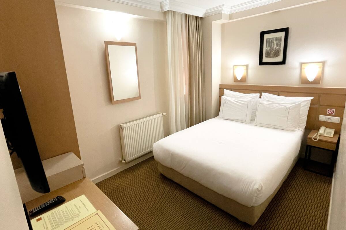 A hotel room with a large bed, a bedside table with a lamp, a framed picture on the wall, and a window with curtains.