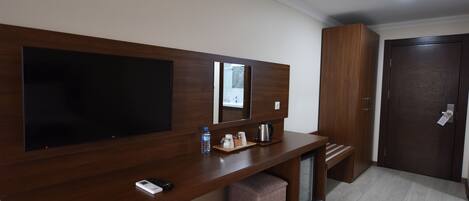 A modern hotel room with a wooden desk, a flat-screen TV, a mirror, a coffee maker, and a small ottoman.