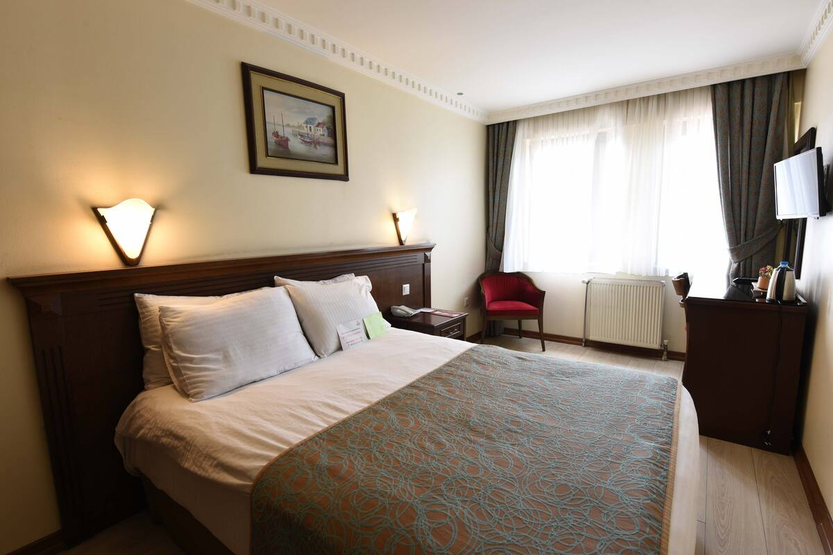 Habitación de hotel con una cama grande, una mesita de noche, una silla roja, un televisor y una ventana con cortinas.