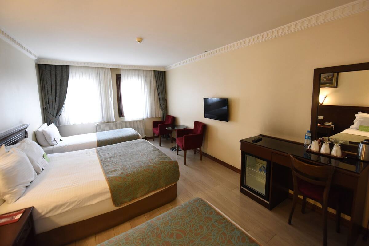 Habitación de hotel con dos camas, un escritorio, una silla, un televisor y un ventanal con cortinas.
