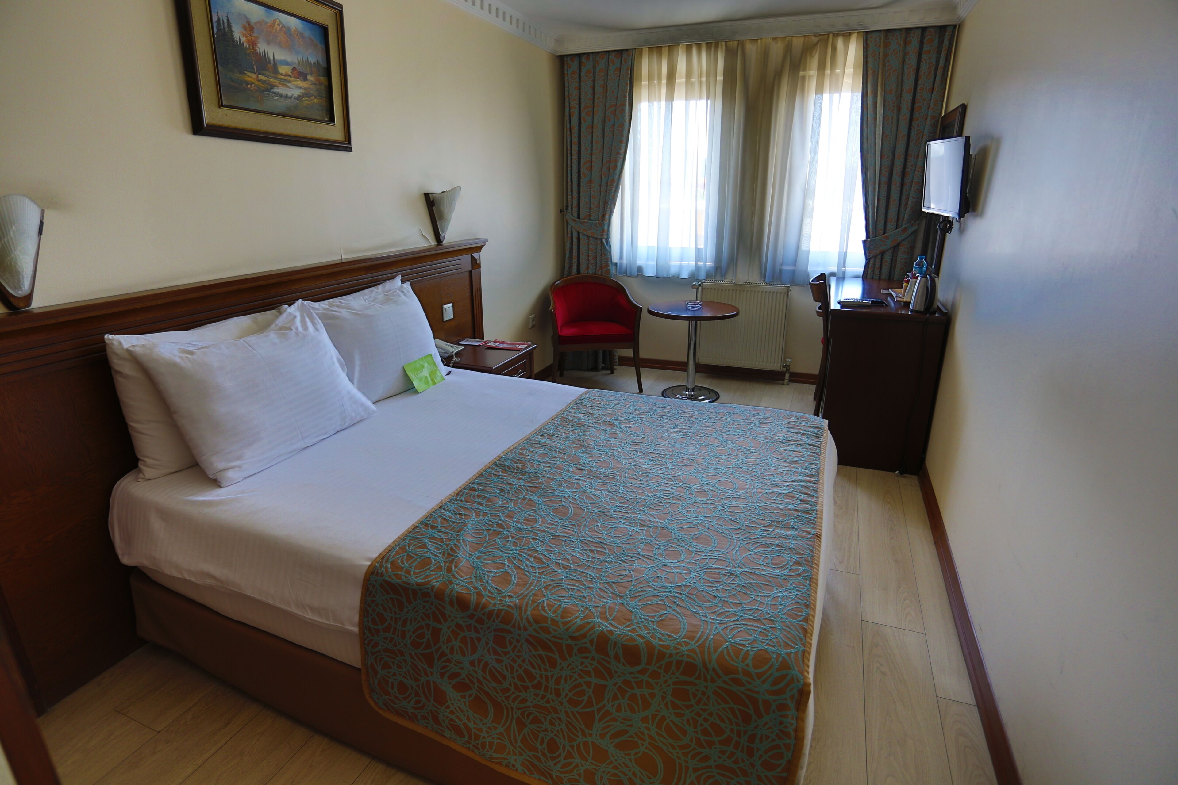 A hotel room with a bed, a desk with a chair, a TV, and a window with curtains.