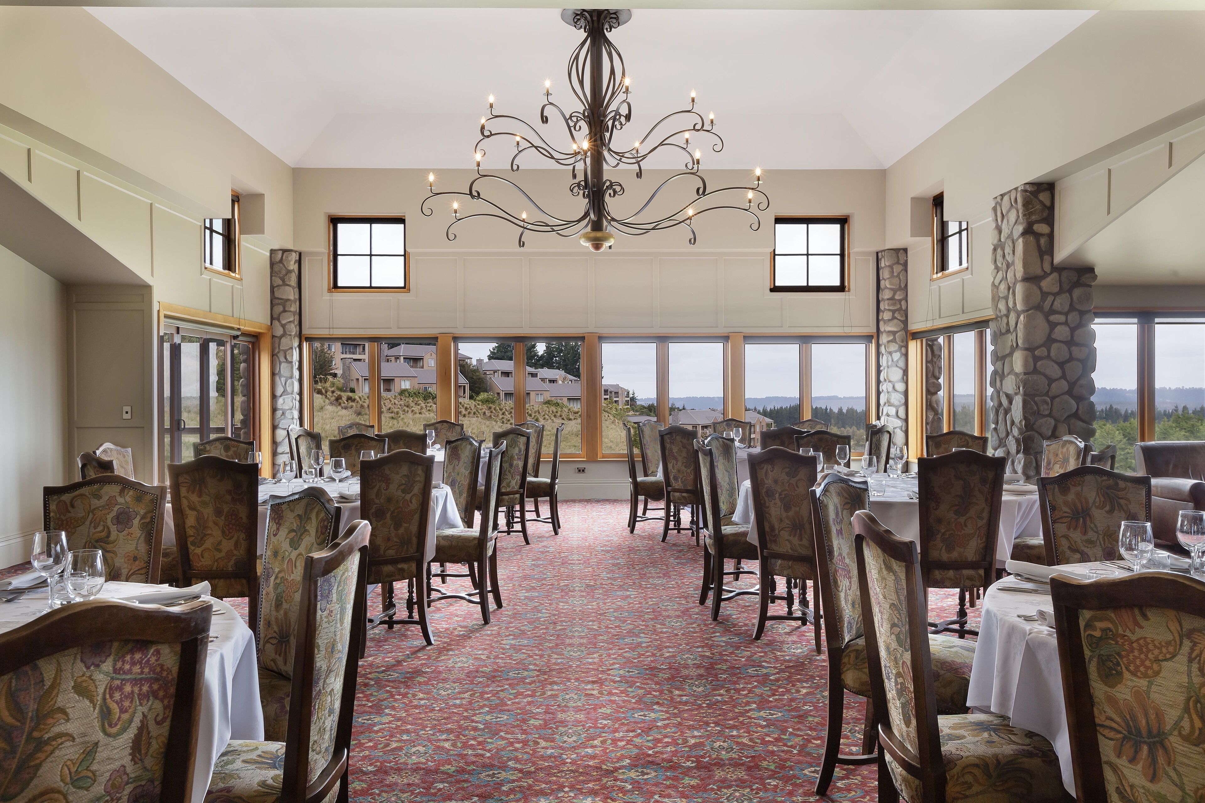 Un comedor con mesas dispuestas para una comida, sillas, una araña y grandes ventanales que ofrecen vistas del paisaje.