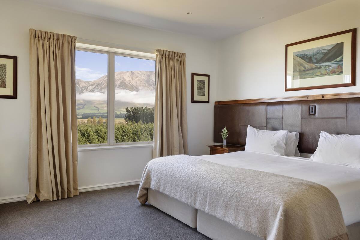 A hotel room with a large bed, a view of mountains through a window, and a framed picture on the wall.