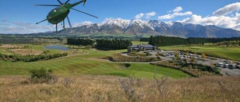 A helicopter flying over a landscape with mountains, a golf course, and a building.