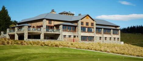A large, multi-story building with a stone facade and multiple windows, situated on a well-maintained lawn with a golf course in the background.
