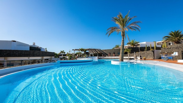 Une grande piscine d’un bleu éclatant, avec un palmier au centre, entourée de bâtiments modernes et d’un mur de pierre.