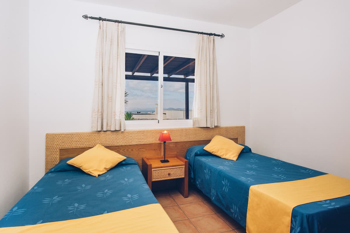 Een kamer met twee bedden, een raam met gordijnen, een nachtkastje met een lamp en uitzicht op zee.