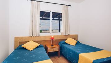 A room with two beds, a window with curtains, a nightstand with a lamp, and a view of the sea.