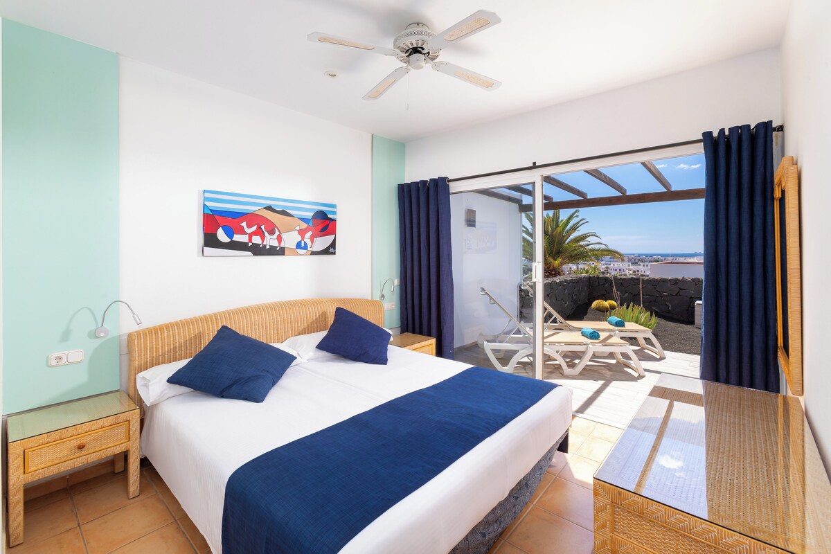 Une chambre à coucher avec un lit, un ventilateur de plafond, un tableau, une table de chevet et une vue sur l’extérieur.