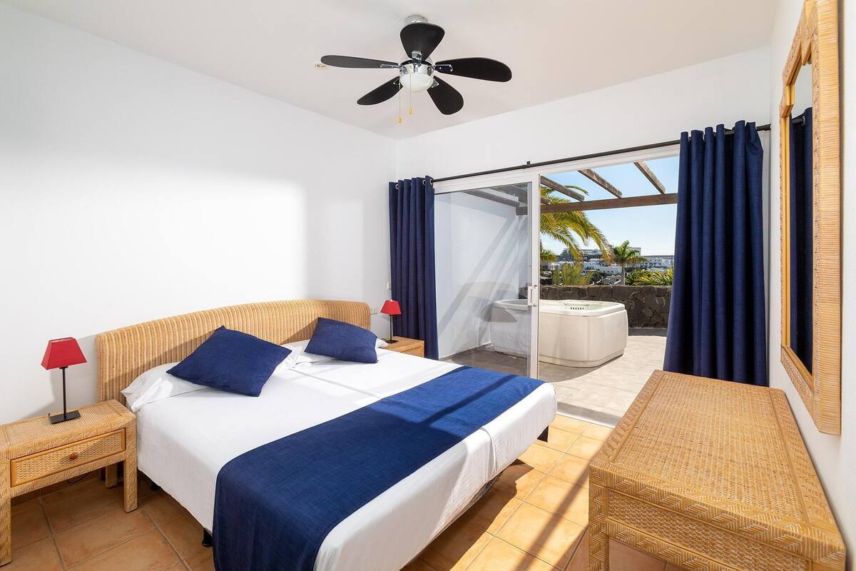 Een hotelkamer met een bed, een plafondventilator, een nachtkastje met lamp, een raam met gordijnen en uitzicht op buiten.