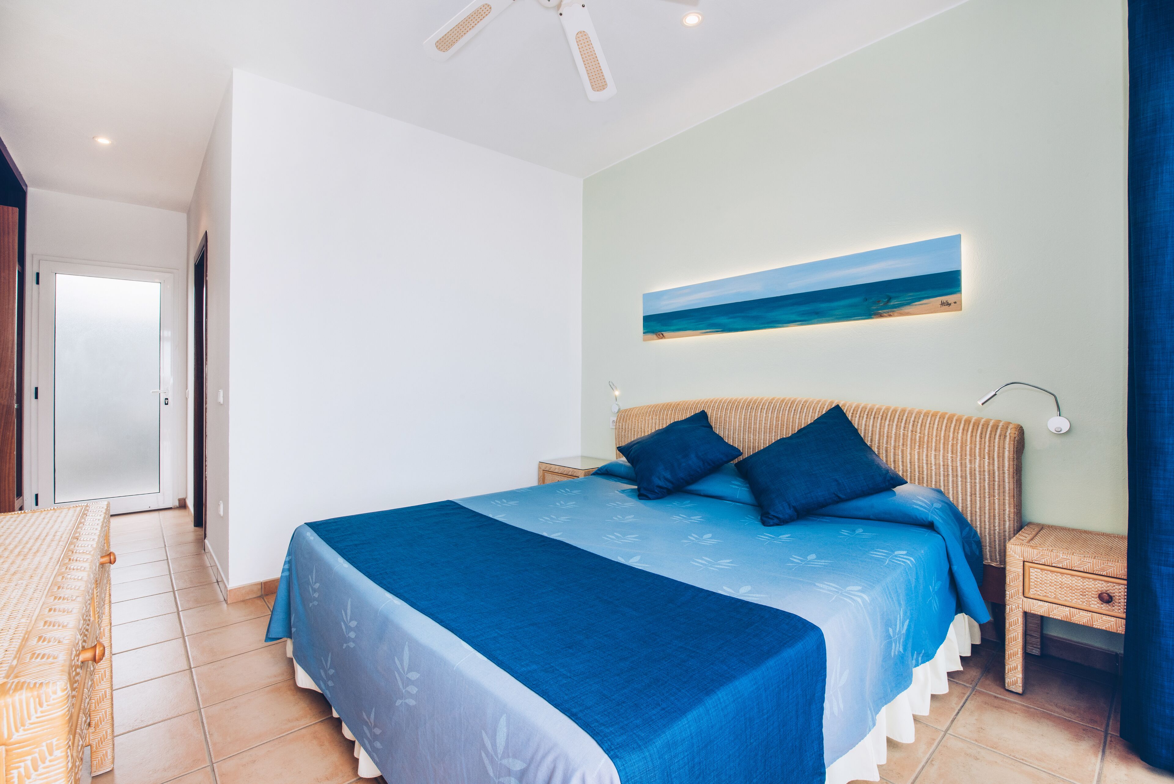 A bedroom with a large bed, a nightstand, a wall-mounted painting, and a door leading to another room.